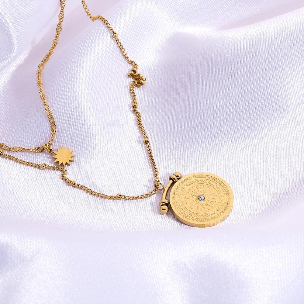 A double layered stainless steel necklace with a gold sun symbol pendant on a shorter chain and a mandala pendant on a longer chain, both made of stainless steel with gold plating.