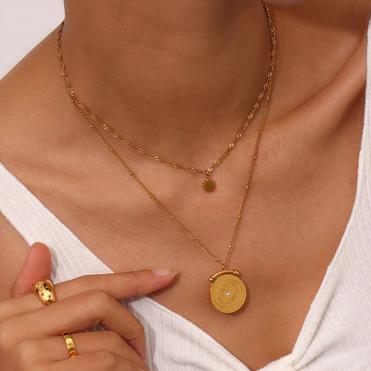 A double layered necklace with a gold-colored sun symbol pendant on a shorter chain and a mandala pendant on a longer chain, both made of stainless steel with gold plating.