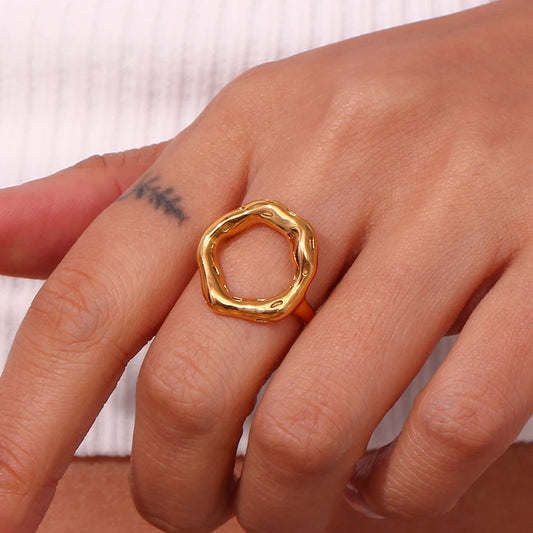 A gold, circle-shaped ring with a textured, asymmetric design, on a model's finger, resembling a stylized representation of life's cycles.