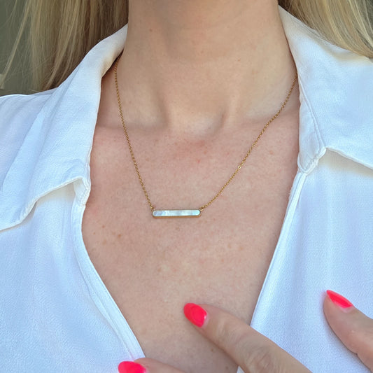 Gold necklace with a bar pendant made of natrual shell, worn by a person in a white shirt.