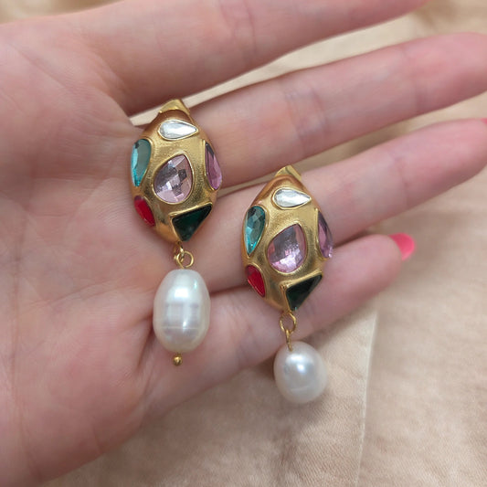 Gold earrings with gemstones and pearls held in a hand against a beige background