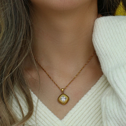 A gold-plated stainless steel necklace with a square pendant featuring a clear crystal, on a model's neck.