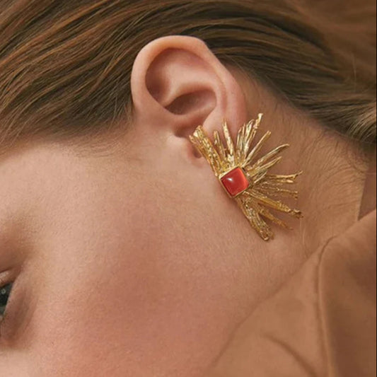 A pair of gold-colored earrings designed to resemble sun rays with a square pink gem at the top on model