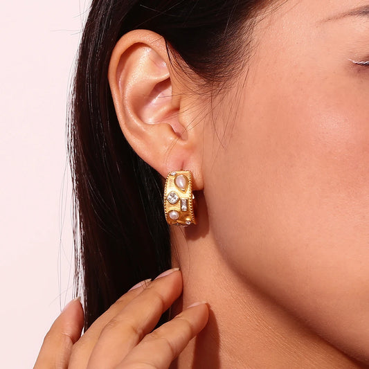 Close-up of a person wearing gold hoop earrings with embedded stones against a light pink background