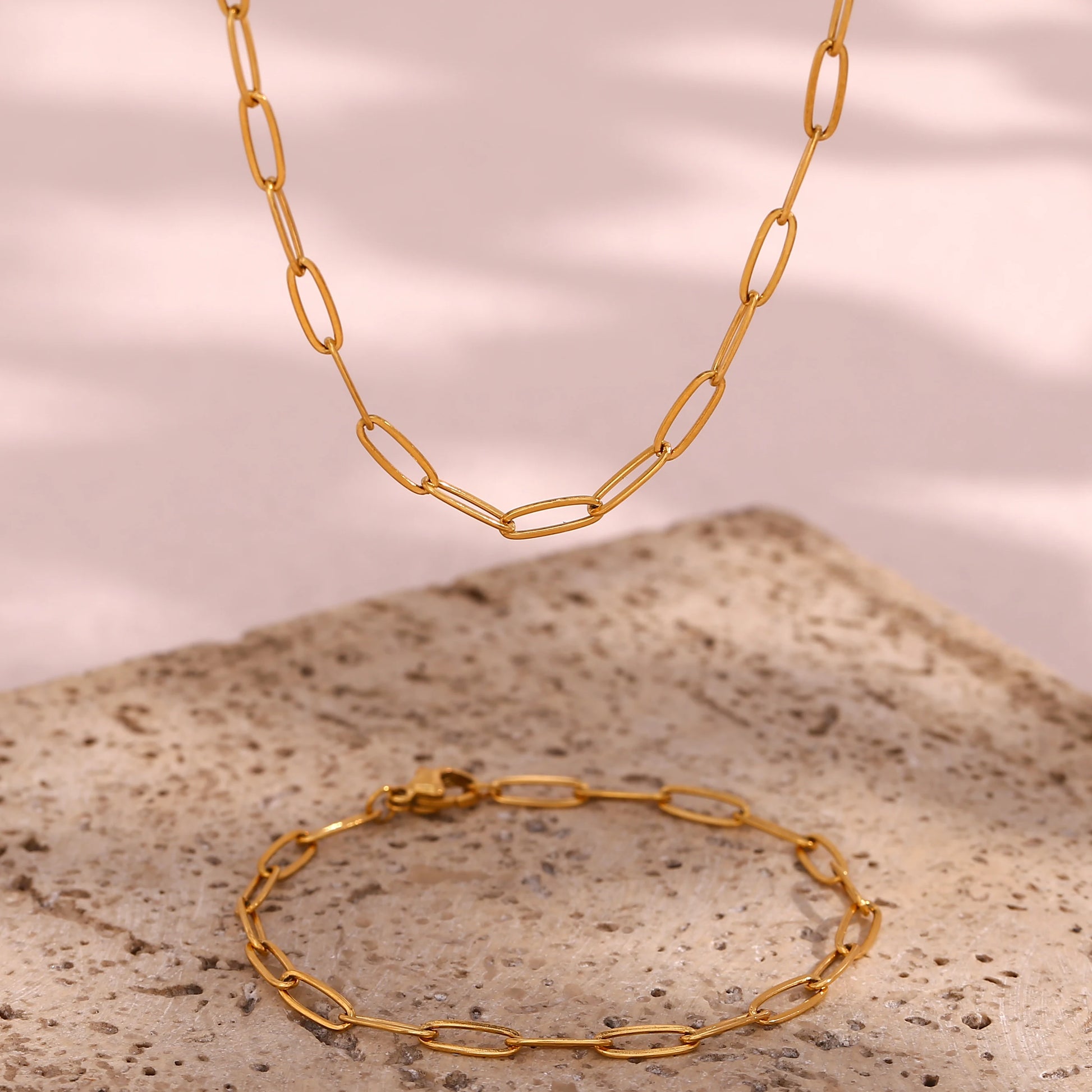 Gold paperclip chain necklace and bracelet on a textured surface with a pinkish-brown background