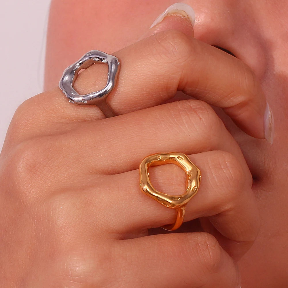 A gold and a silver, circle-shaped ring with a textured, asymmetric design, on a model's finger, resembling a stylized representation of life's cycles.