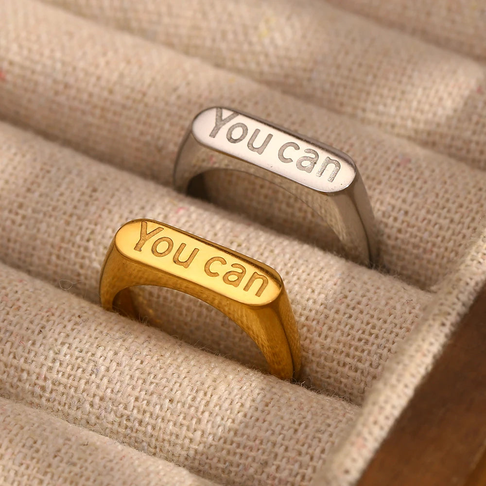 stainless steel signet rings with the word 'you can' engraved on the top. In silver and gold in a jewelry display 