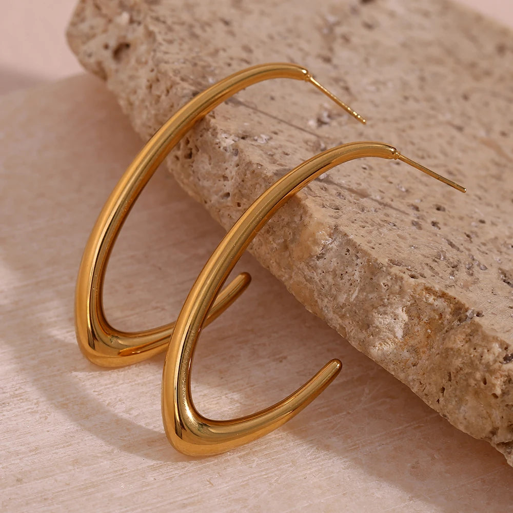 A pair of elegant large Hoop Earrings, presented on a textured stone surface. The earrings are designed in a bold, minimalist style, with a glossy gold finish that catches the light, enhancing their appeal.