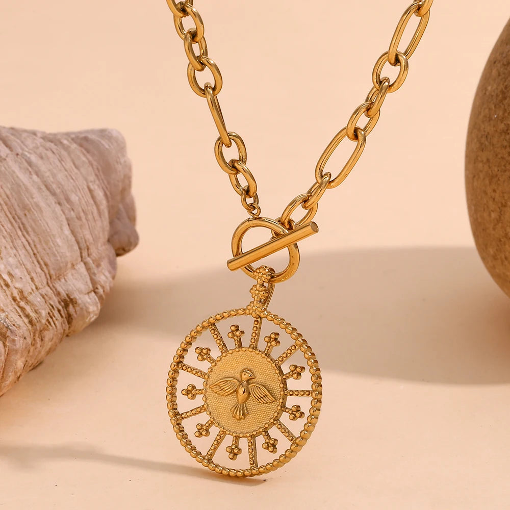 A stainless steel gold chunky necklace with a circular pendant featuring a bird, displayed against a neutral background with a shell to the left.