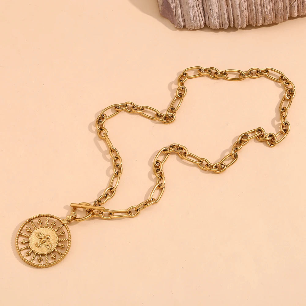 A stainless steel gold chunky necklace with a circular pendant featuring a bird pendant, displayed against a neutral background with a shell to the left.
