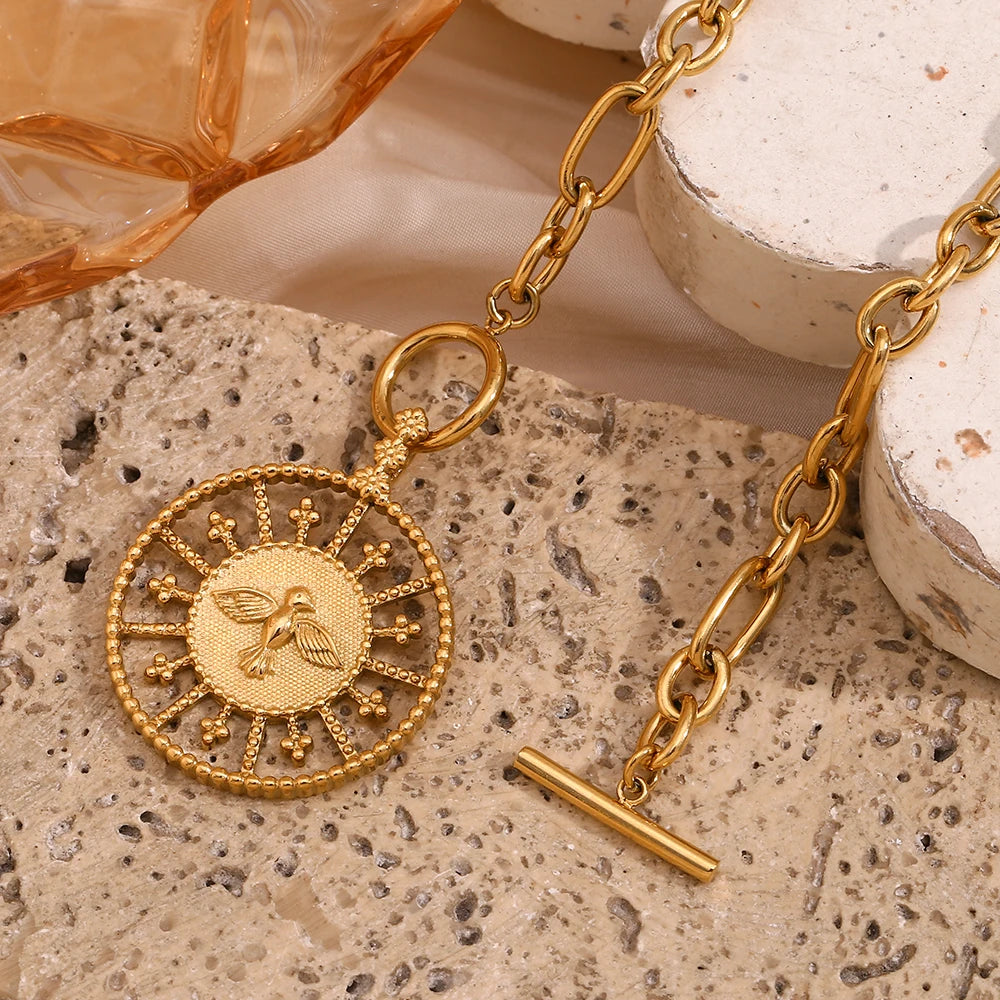 A stainless steel gold chunky necklace with a circular pendant featuring a bird design, displayed against a neutral background with a shell to the left.