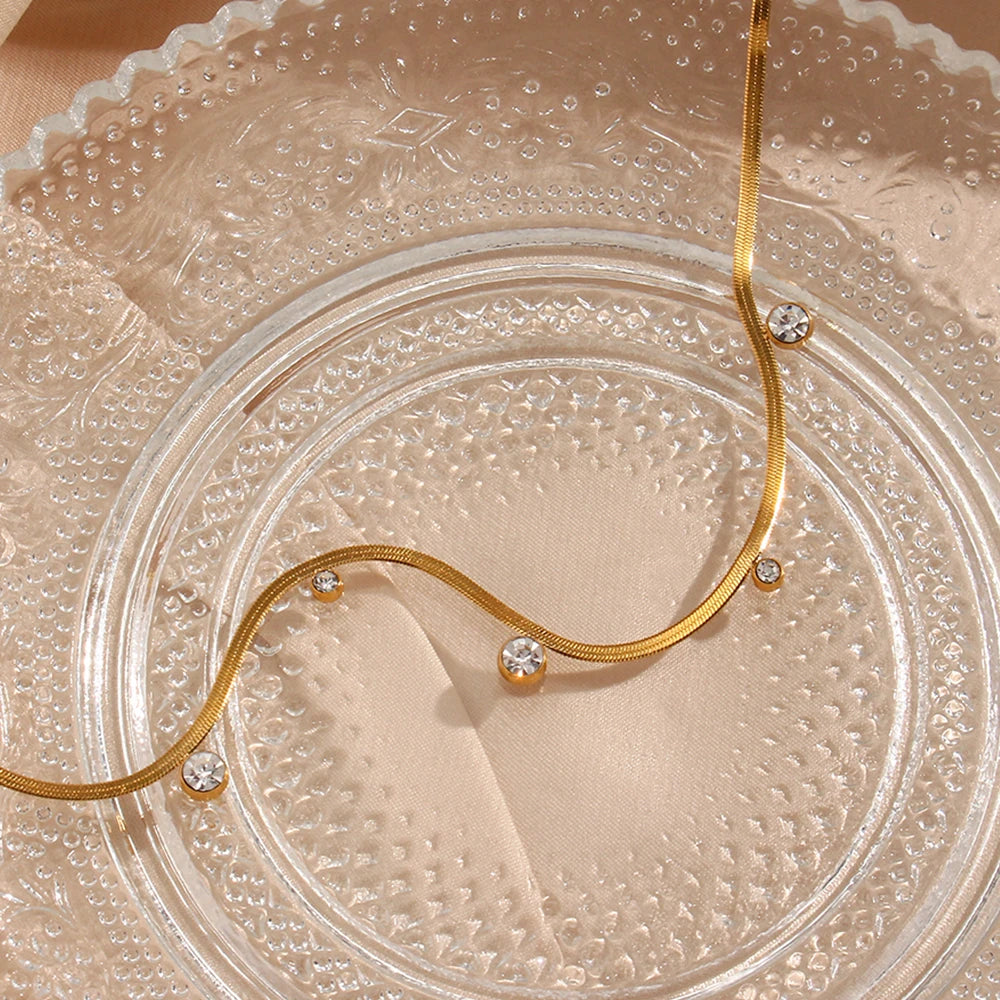 A 18K gold-plated stainless steel necklace with round zirconia crystals, on a glass textured dish.
