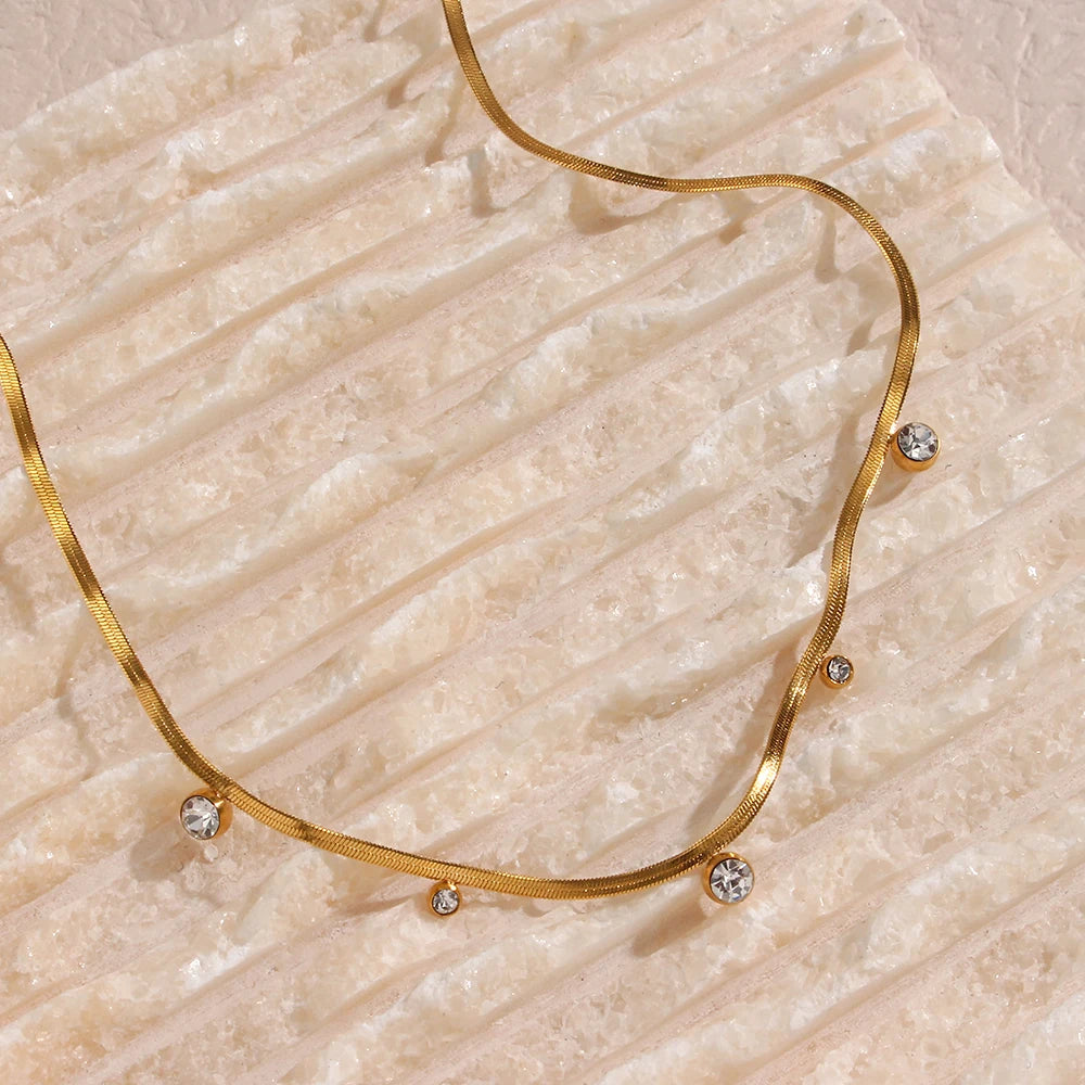A 18K gold-plated stainless steel necklace with round zirconia crystals, on a beige textured background.