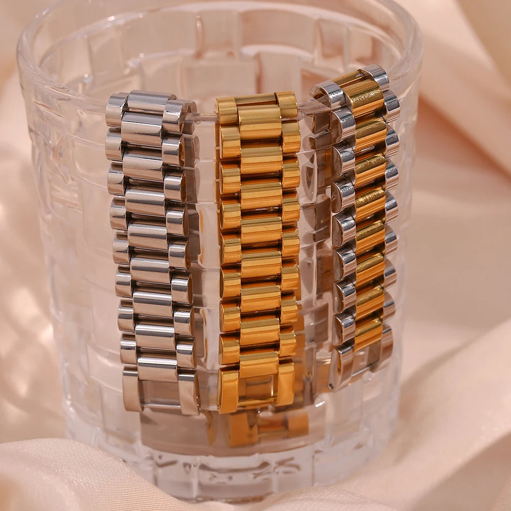 Two metallic bracelets, one silver and one gold, displayed over the edge of a glass against a soft pink background.