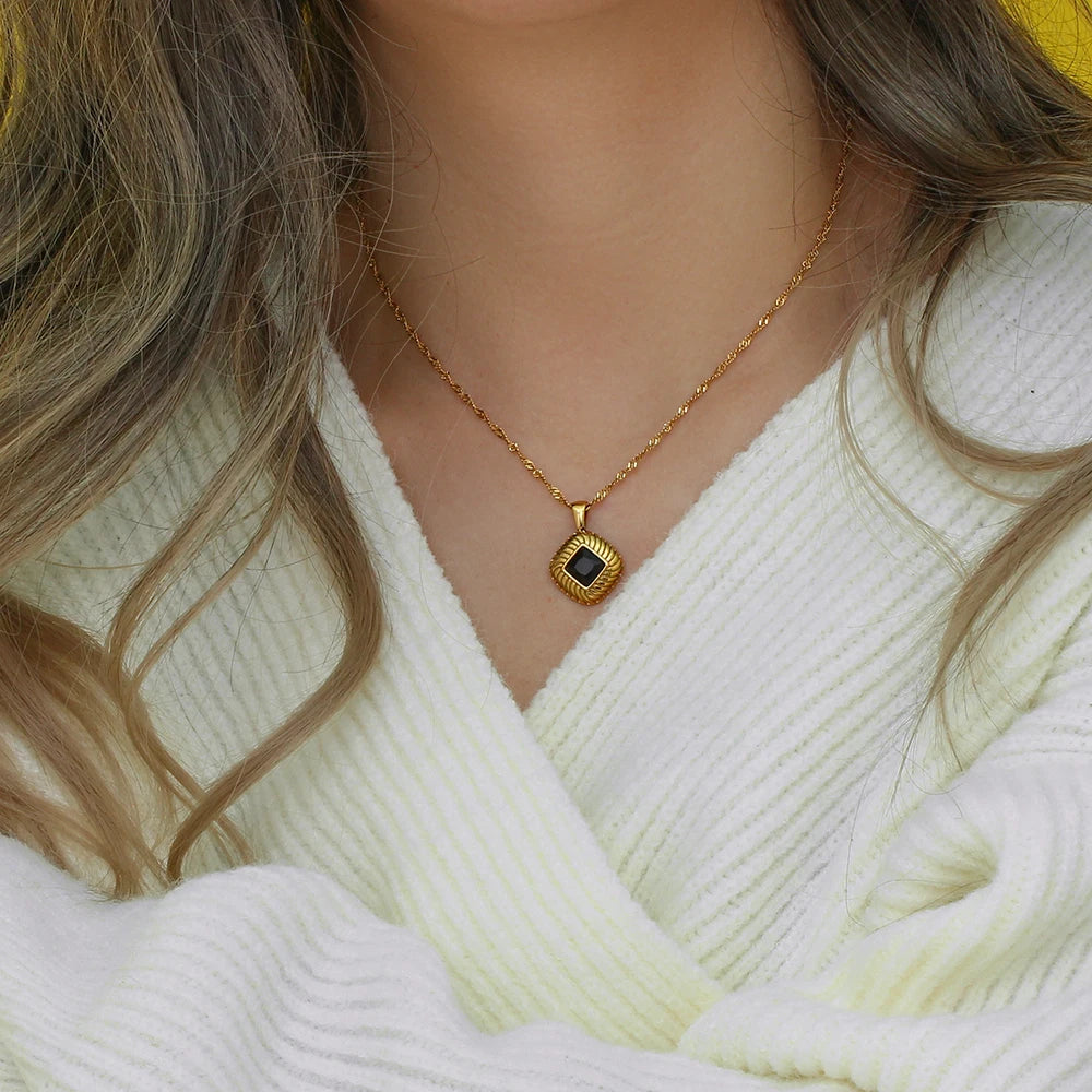 A gold stainless steel necklace with a square pendant featuring a black crystal, on a model's neck.