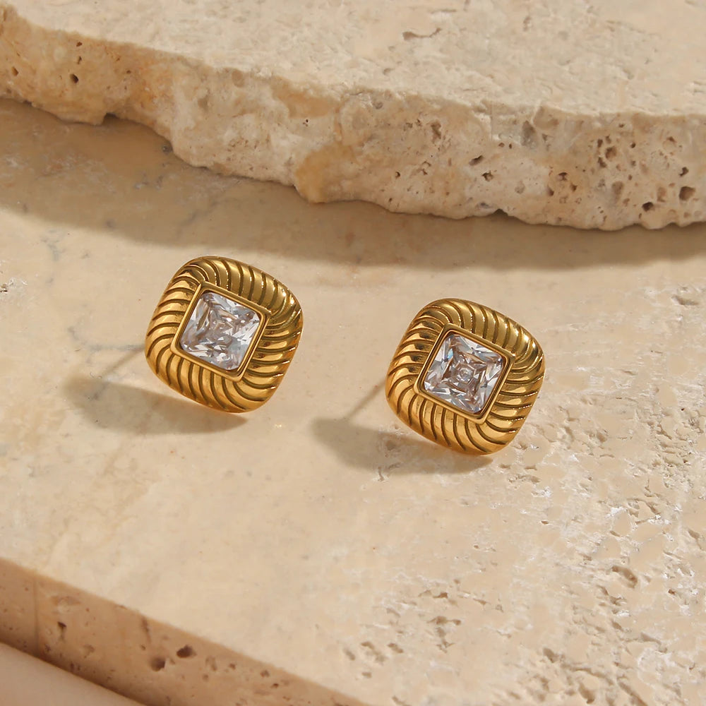 A pair of stainless steel stud earrings with a geometric design and a central clear crystal, displayed on a textured beige tile.