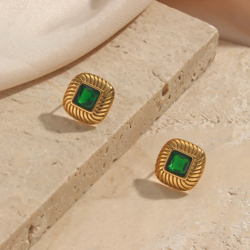 A pair of stainless steel stud earrings with a geometric design and a central green crystal, displayed on a textured beige tile.