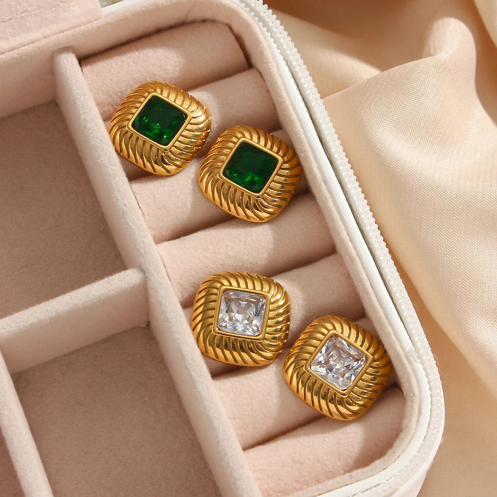 two pairs of stainless steel stud earrings with a geometric design and a central crystal, displayed on a beige jewelry box.