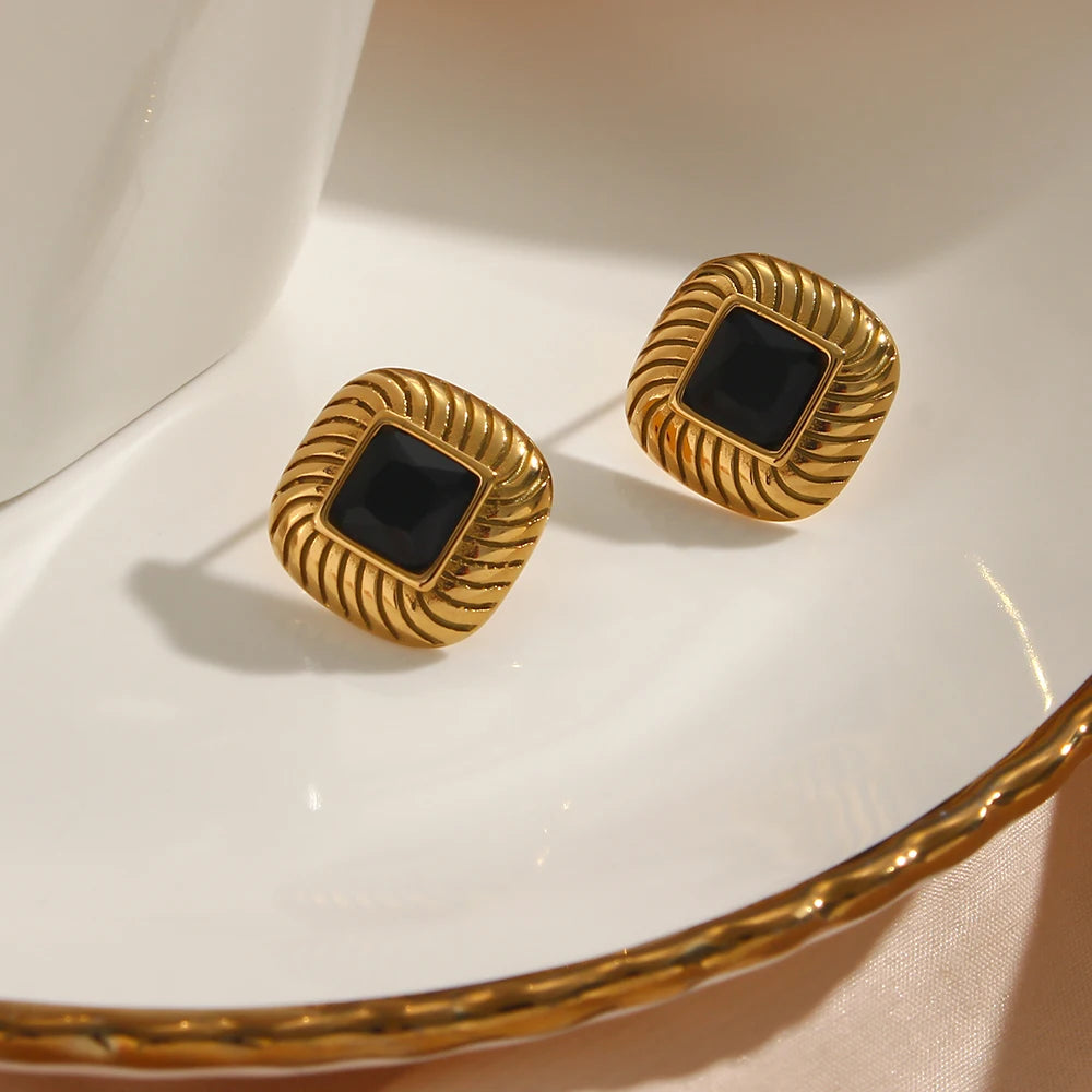 A pair of stainless steel stud earrings with a geometric design and a central black crystal, displayed on a textured beige tile.