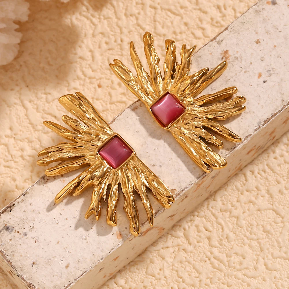 A pair of gold-colored earrings designed to resemble sun rays with a square pink gem at the top flat lay
