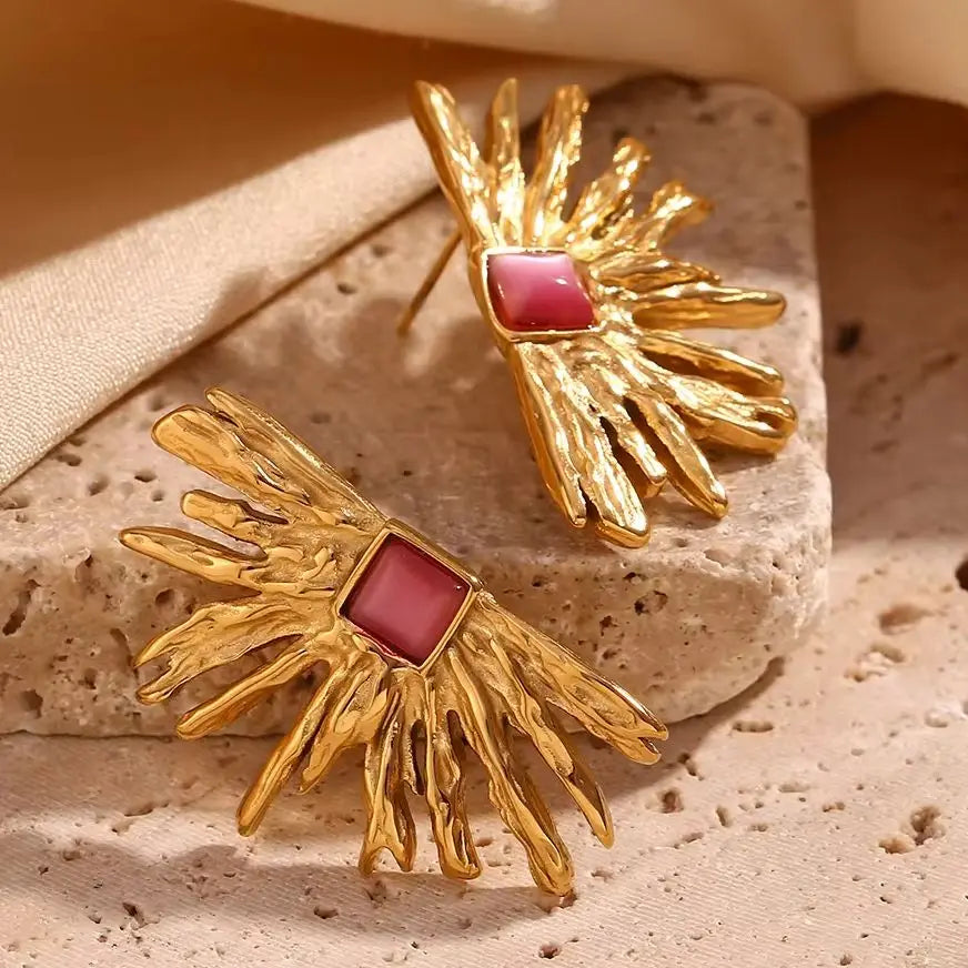 A pair of gold-colored earrings designed to resemble sun rays with a square pink gem at the top closeup