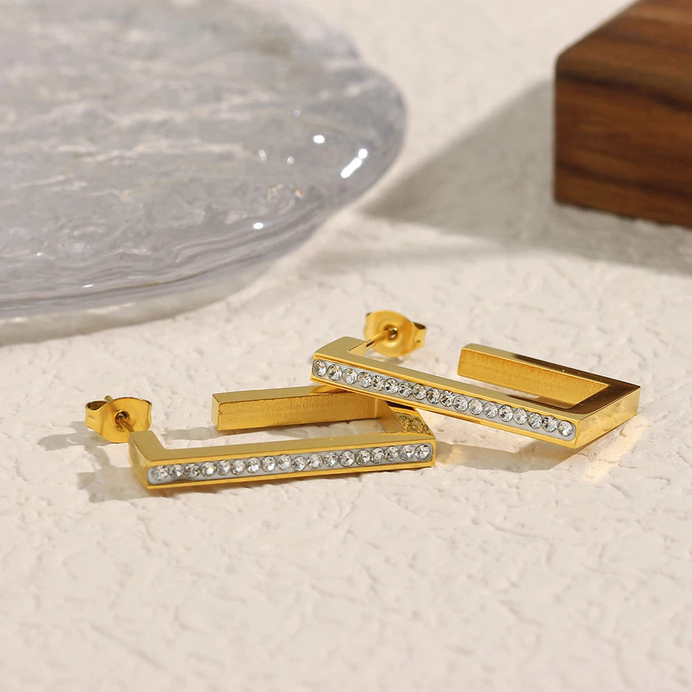 Gold rectangular earrings with clear stones on a textured surface