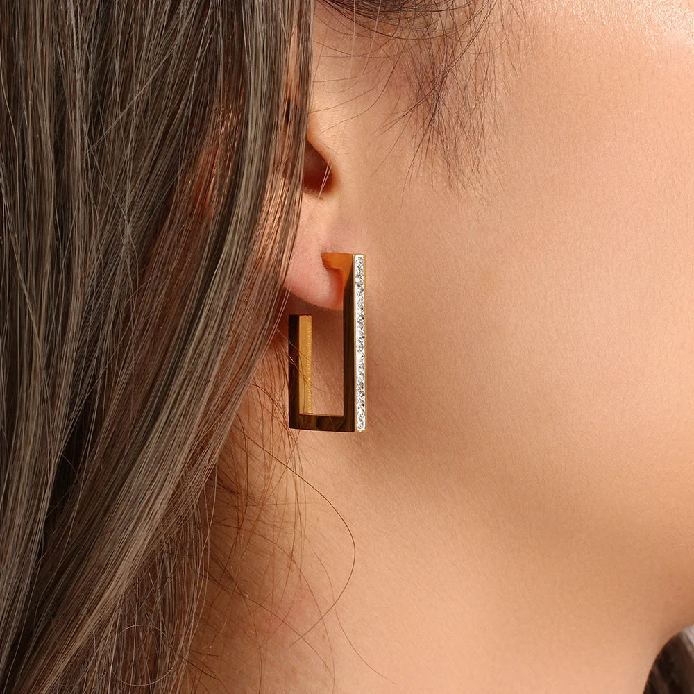 Close-up of a person wearing a gold rectangular hoop earring with small diamonds.